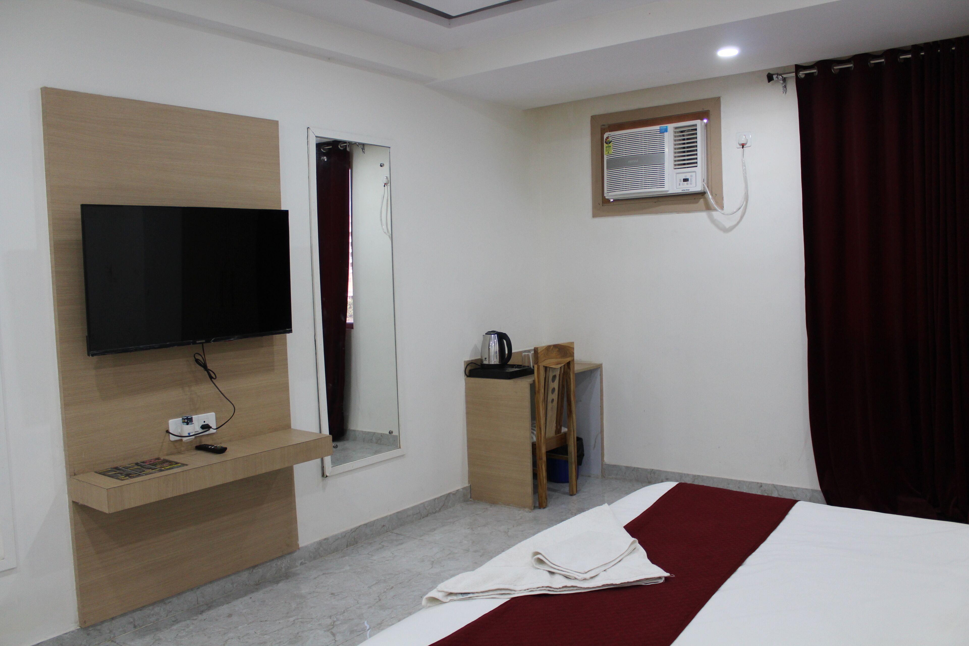 A hotel room with a bed, a TV mounted on a wooden panel, a small window with an air conditioning unit, and a desk with a chair.