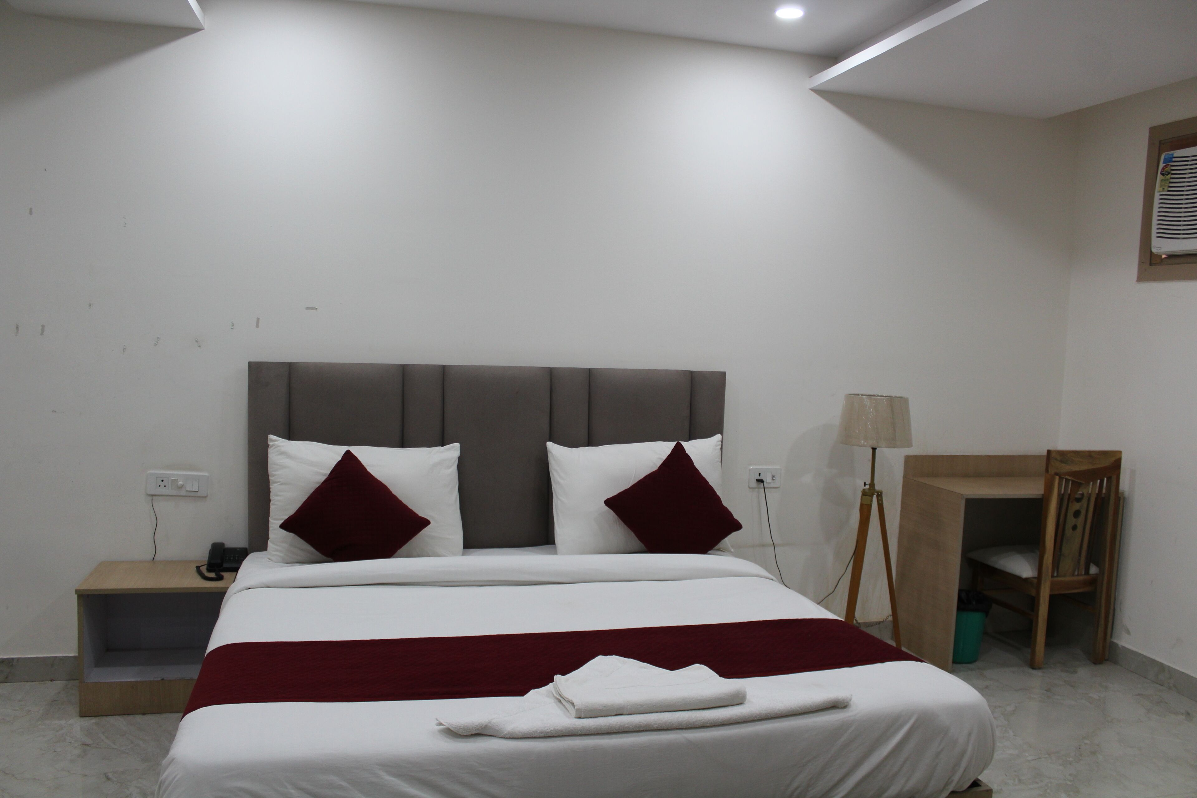 A neatly made bed with white and red bedding, a bedside table with a lamp, and a small wooden desk with a chair.