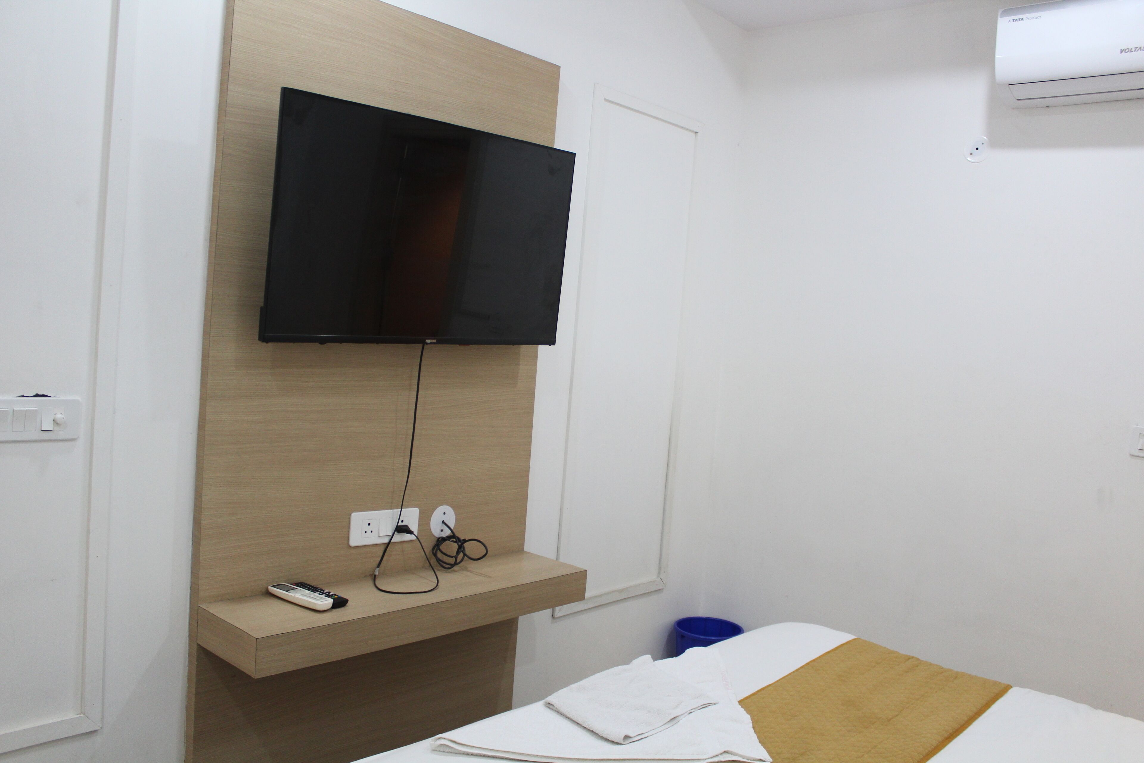 A hotel room with a bed, a TV mounted on the wall, a bedside table with a remote control, and a blue trash bin.