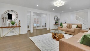 A spacious living room with a beige sofa, a glass coffee table, and a wooden side table. There is a staircase leading to another level, and a large mirror on the wall. - Luxury 7BR Buckhead Oasis w Pool Gameroom Firepit (Atlanta)