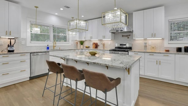 A modern kitchen with white cabinetry, a marble island with bar stools, pendant lighting, and a stainless steel range hood. - Luxury 7BR Buckhead Oasis w Pool Gameroom Firepit (Atlanta)