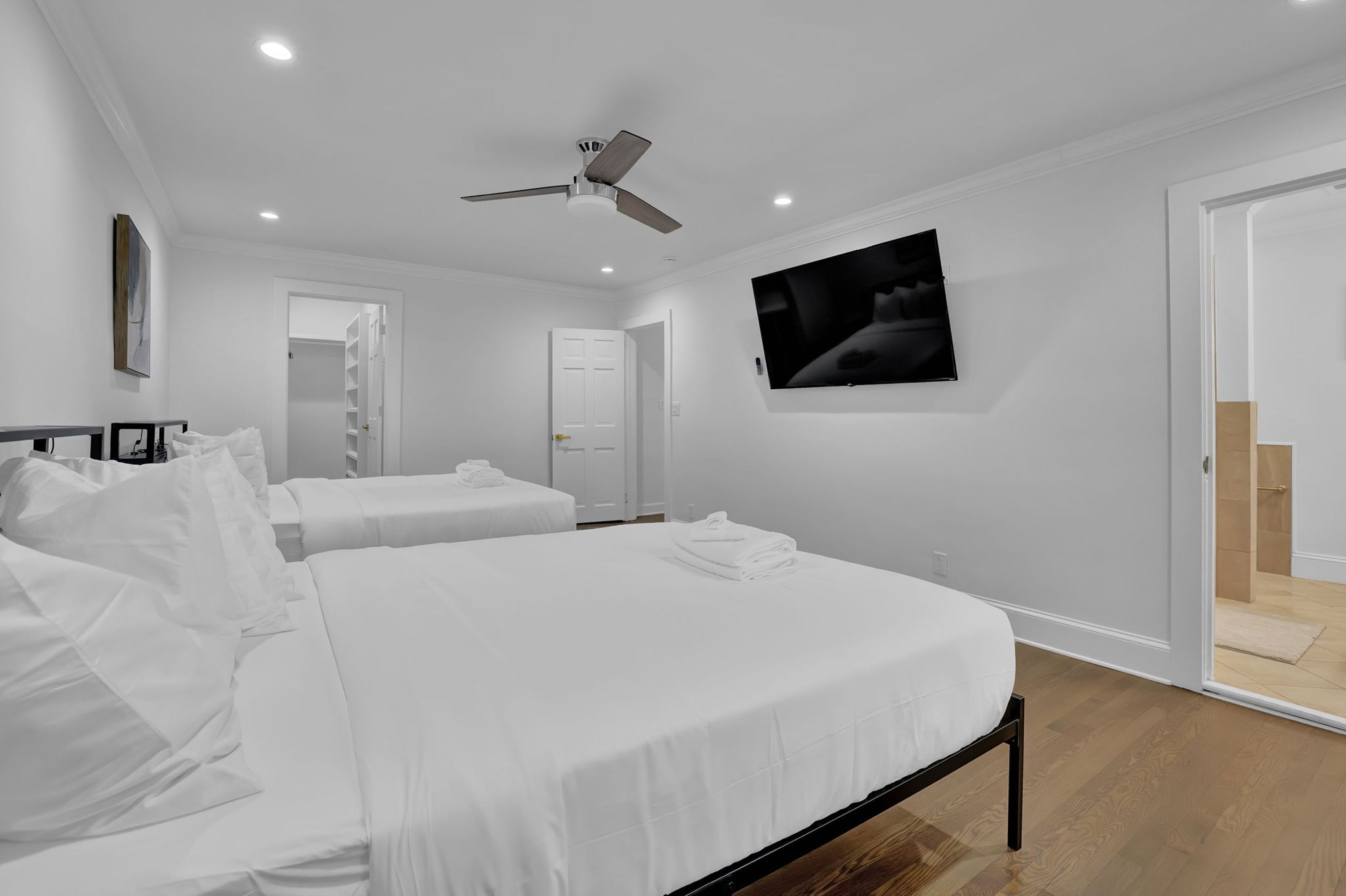 A hotel room with two beds, a ceiling fan, recessed lighting, a TV on the wall, and a door leading to a shower area.