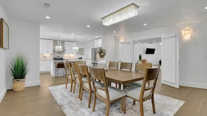 A modern dining area with a wooden table and chairs, a chandelier, and a kitchen in the background. - Luxury 7BR Buckhead Oasis w Pool Gameroom Firepit (Atlanta)