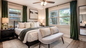 A bedroom with a bed, a bench, a nightstand, a ceiling fan, and two windows with curtains.
