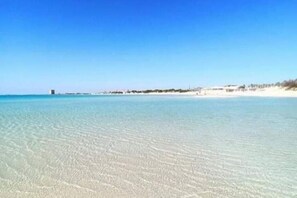 A beach with clear shallow water, distant buildings, and a sandy shore.