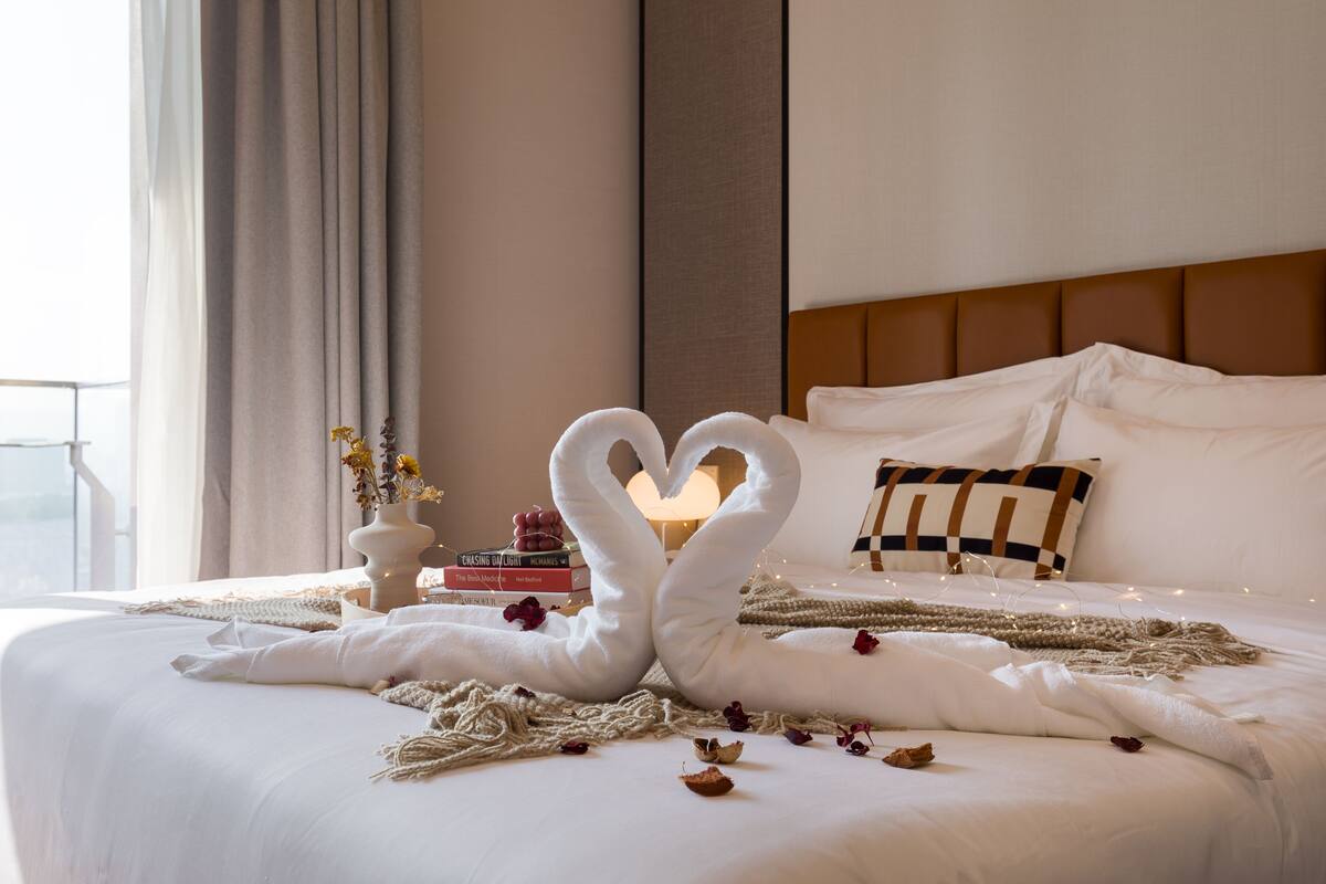 A neatly made bed with a heart-shaped towel arrangement, a bedside table with books, and a lamp.