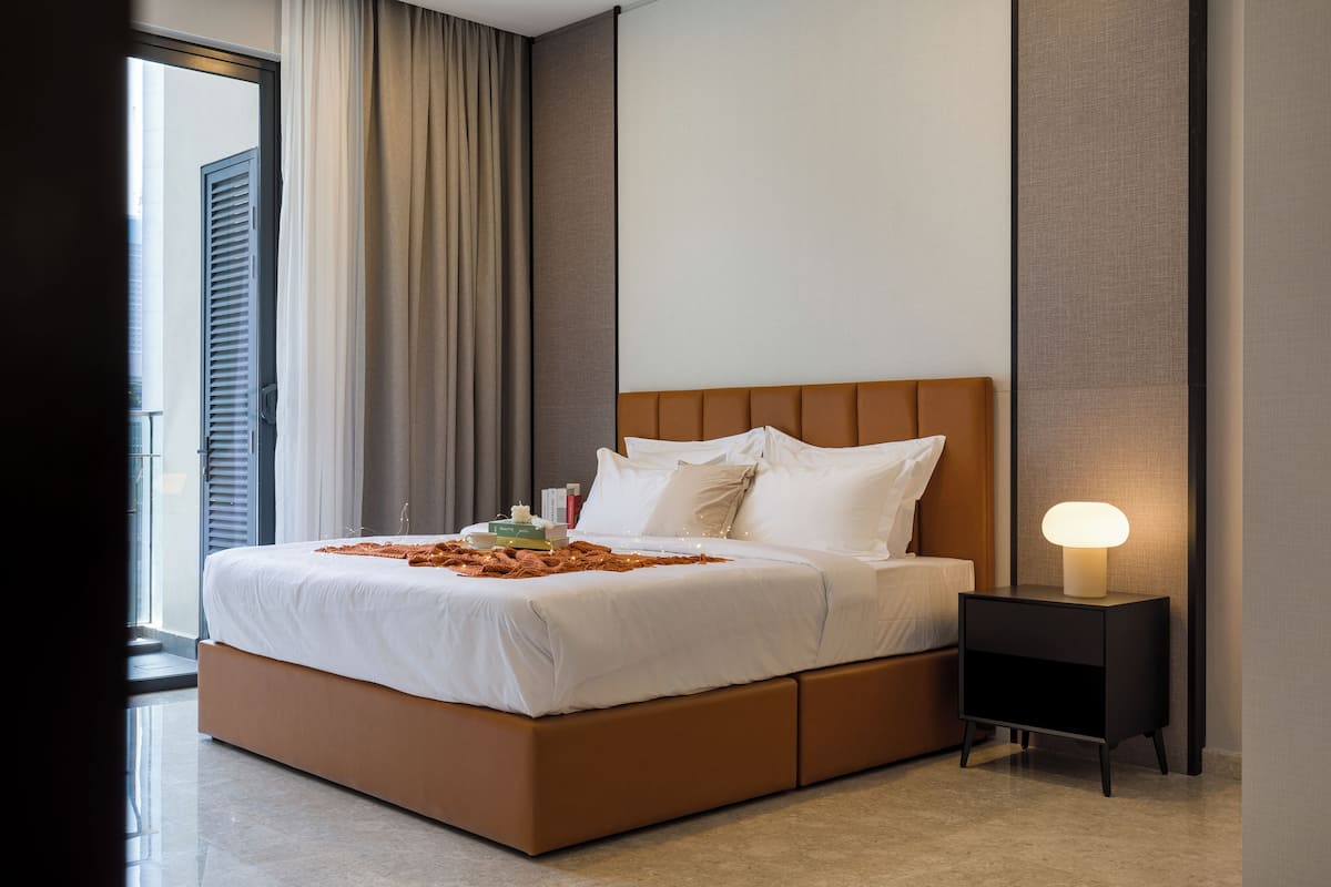 A modern hotel room with a large bed, a nightstand with a lamp, and a view of the city through the window.