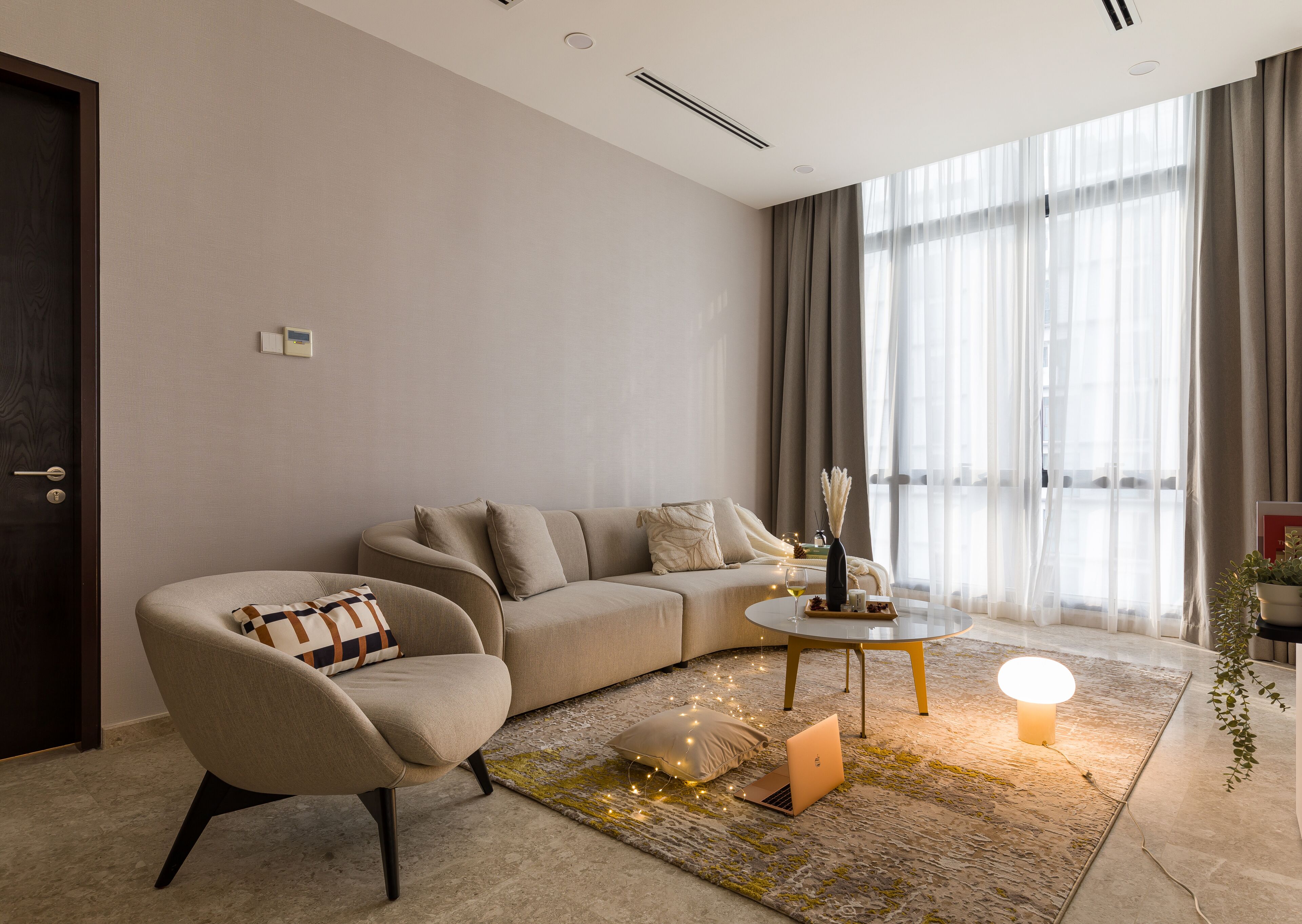 Una sala de estar moderna con un sofá, un sillón y una mesa de centro. Hay un ordenador portátil en el suelo, una lámpara en la mesa y una planta al lado.