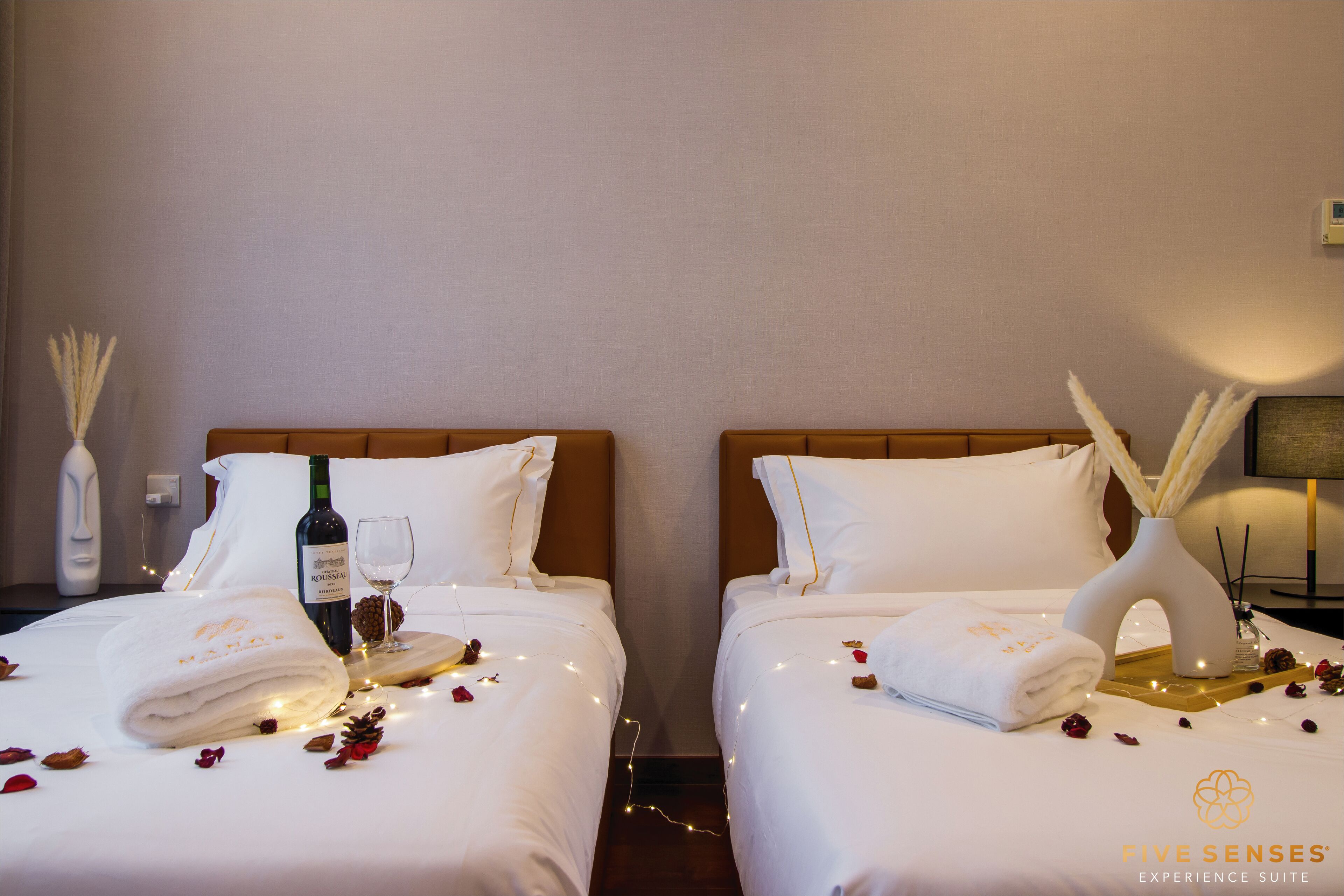 A hotel room with two beds, each with white linens, a bottle of wine, a glass, and decorative items like dried flowers and candles.