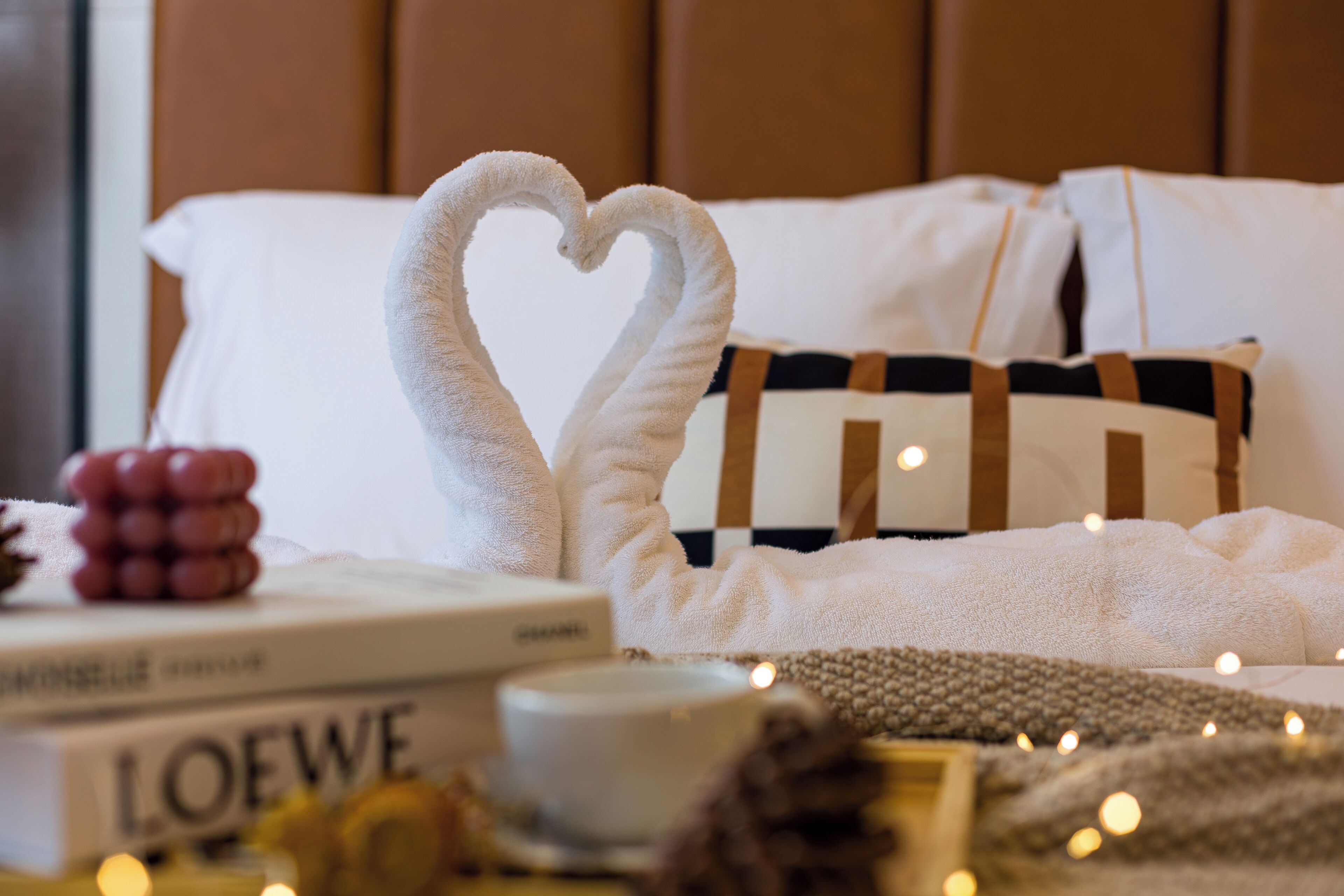 Een net opgemaakt bed met decoratieve handdoek in de vorm van een hart, een boek getiteld 'LOEWE' en een kleine kaars op de nachtkastje.