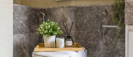 Un baño con pared de mármol, ducha, planta en maceta, vela y bandeja con un difusor de aroma de caña.