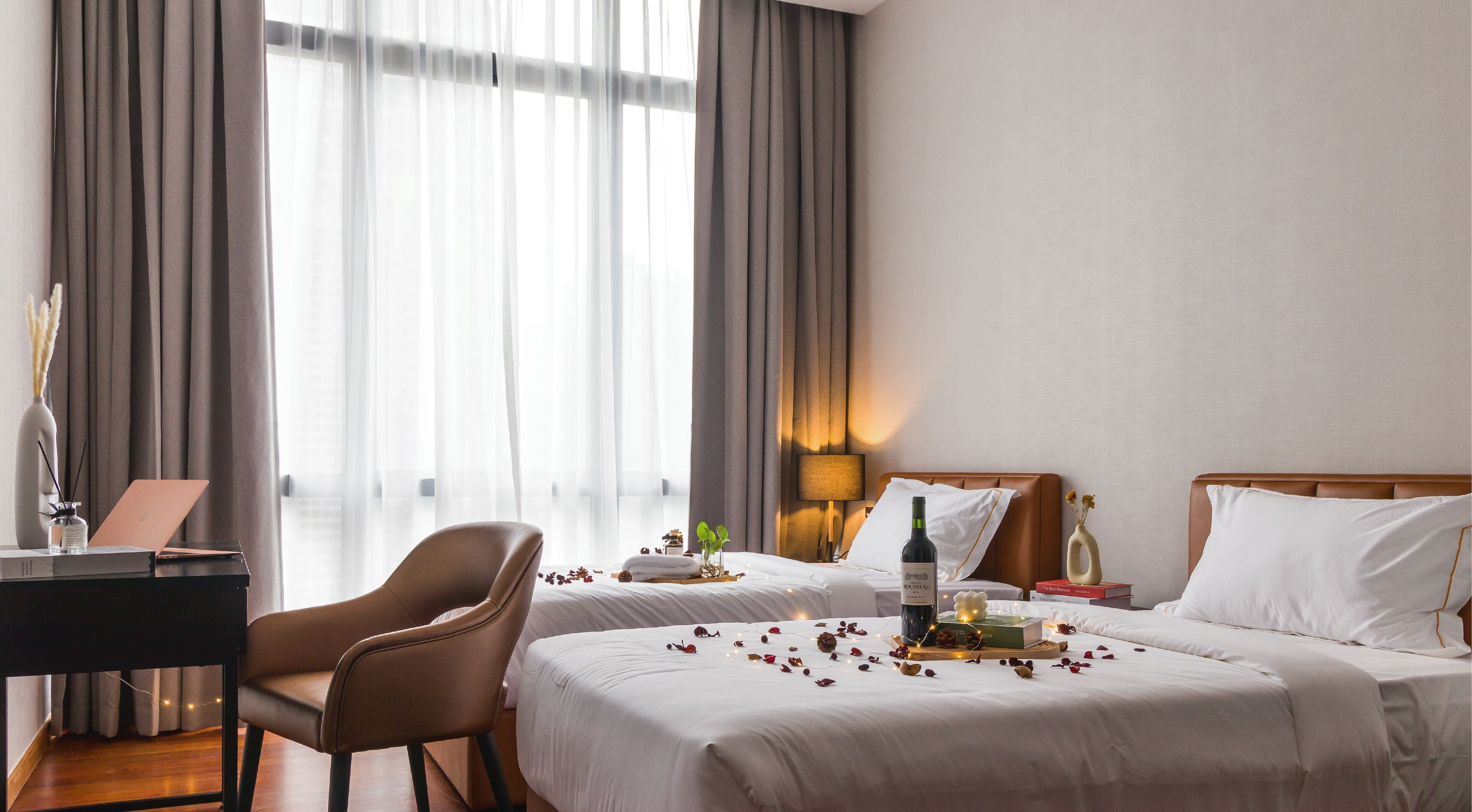 A hotel room with two beds, a desk with a laptop, a chair, a bedside table with a bottle and a book, and a lamp.
