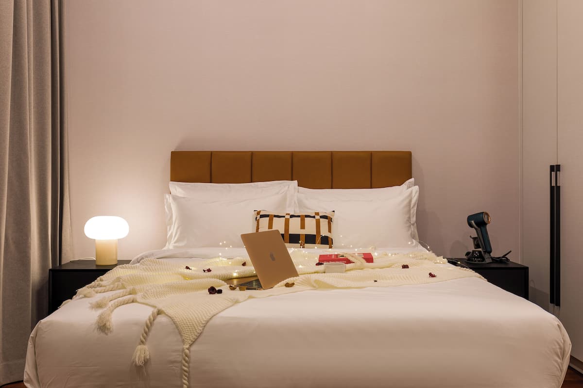 A neatly made bed with a laptop, a bedside lamp, and a small table with a decorative item.