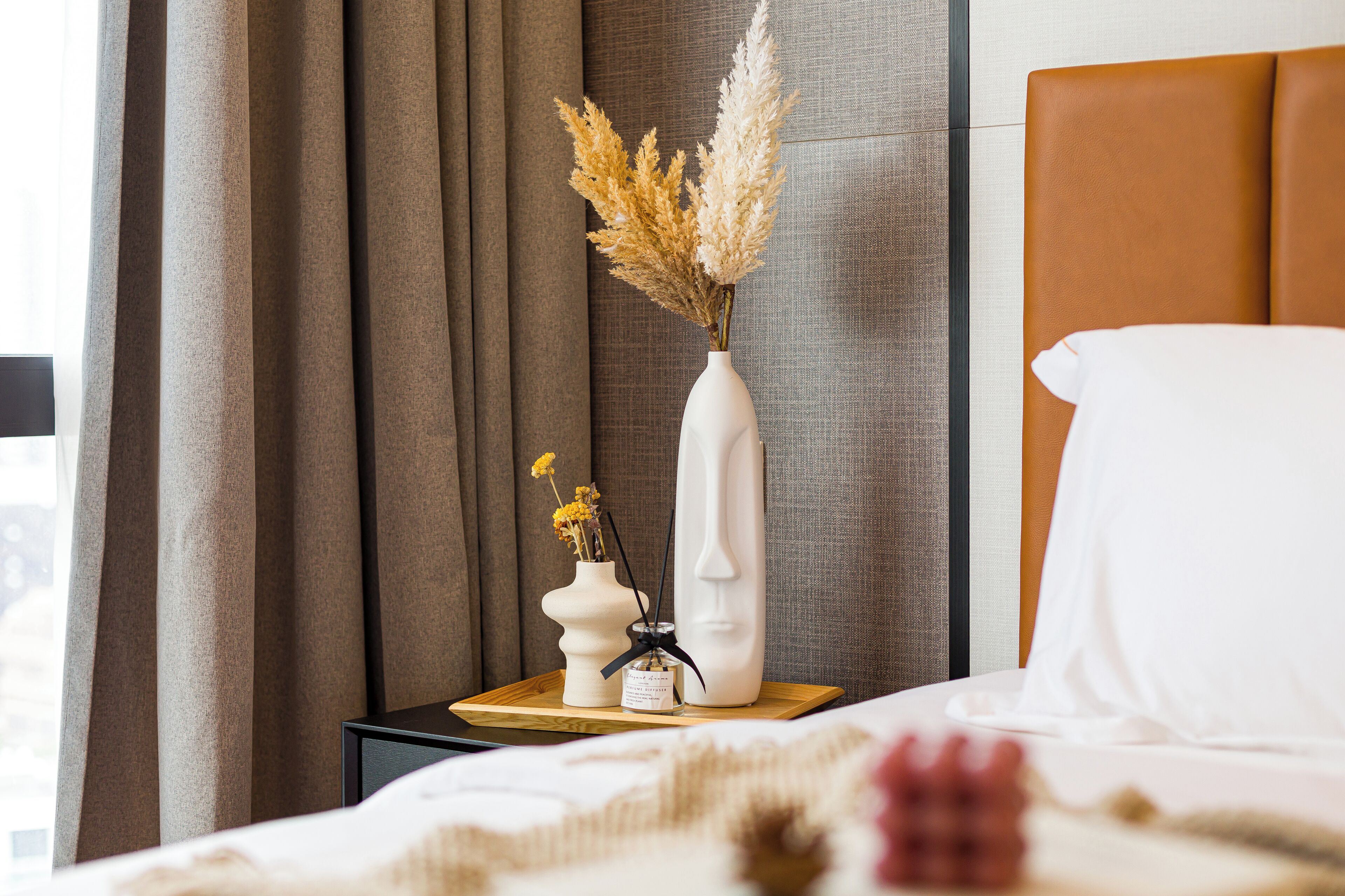 Una cama bien hecha con sábanas blancas, un jarrón blanco con flores secas y un pequeño objeto decorativo sobre una mesita de noche de madera.