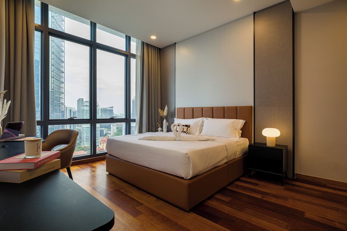 A modern hotel room with a large bed, a desk with a laptop, and a view of the city through large windows.