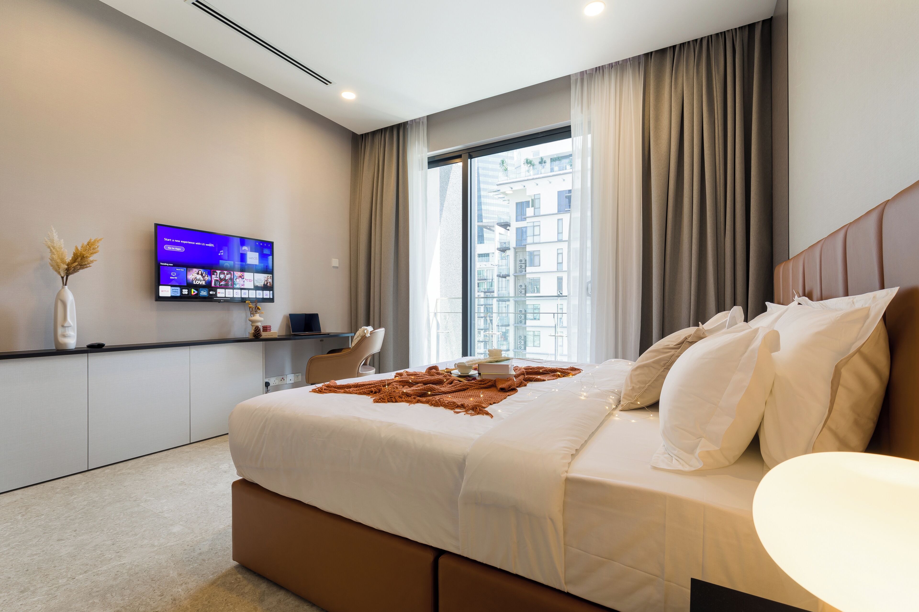 A modern hotel room with a large bed, a flat-screen TV mounted on the wall, and a view of the city through the window.