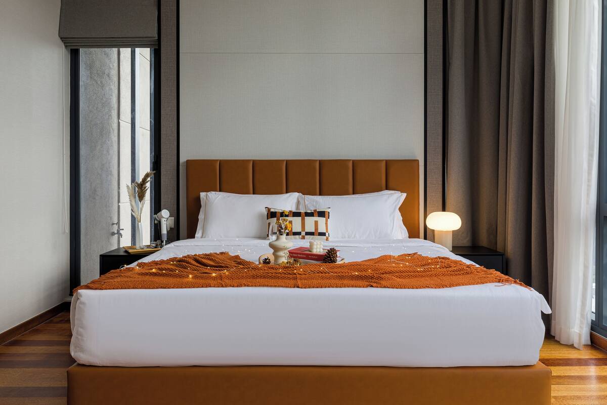 A neatly made bed with white linens and an orange throw blanket, a bedside table with a lamp, and a window with curtains.