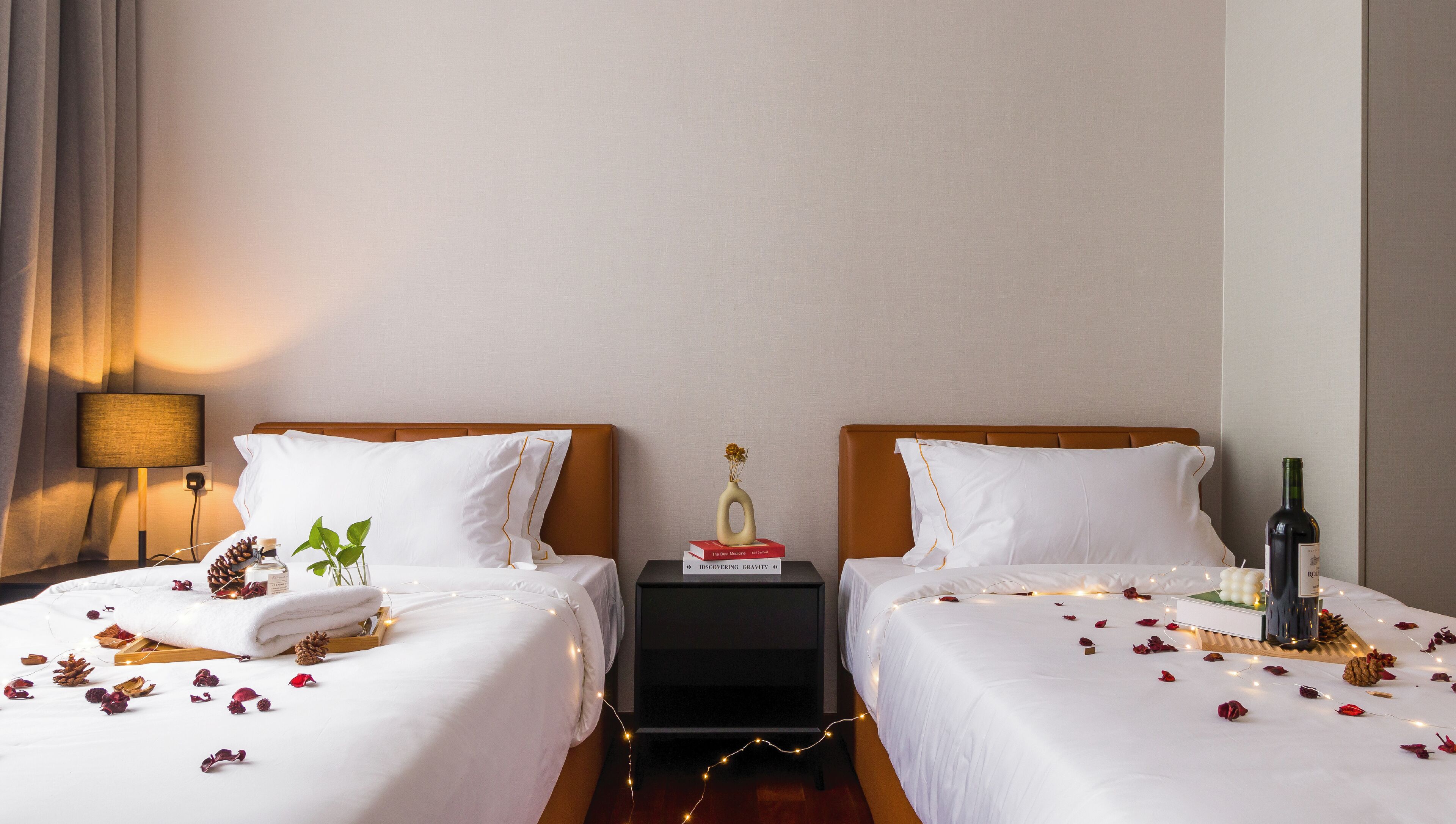 Une chambre d’hôtel avec deux lits, une table de chevet avec une lampe, un vase de fleurs et une bouteille de vin.