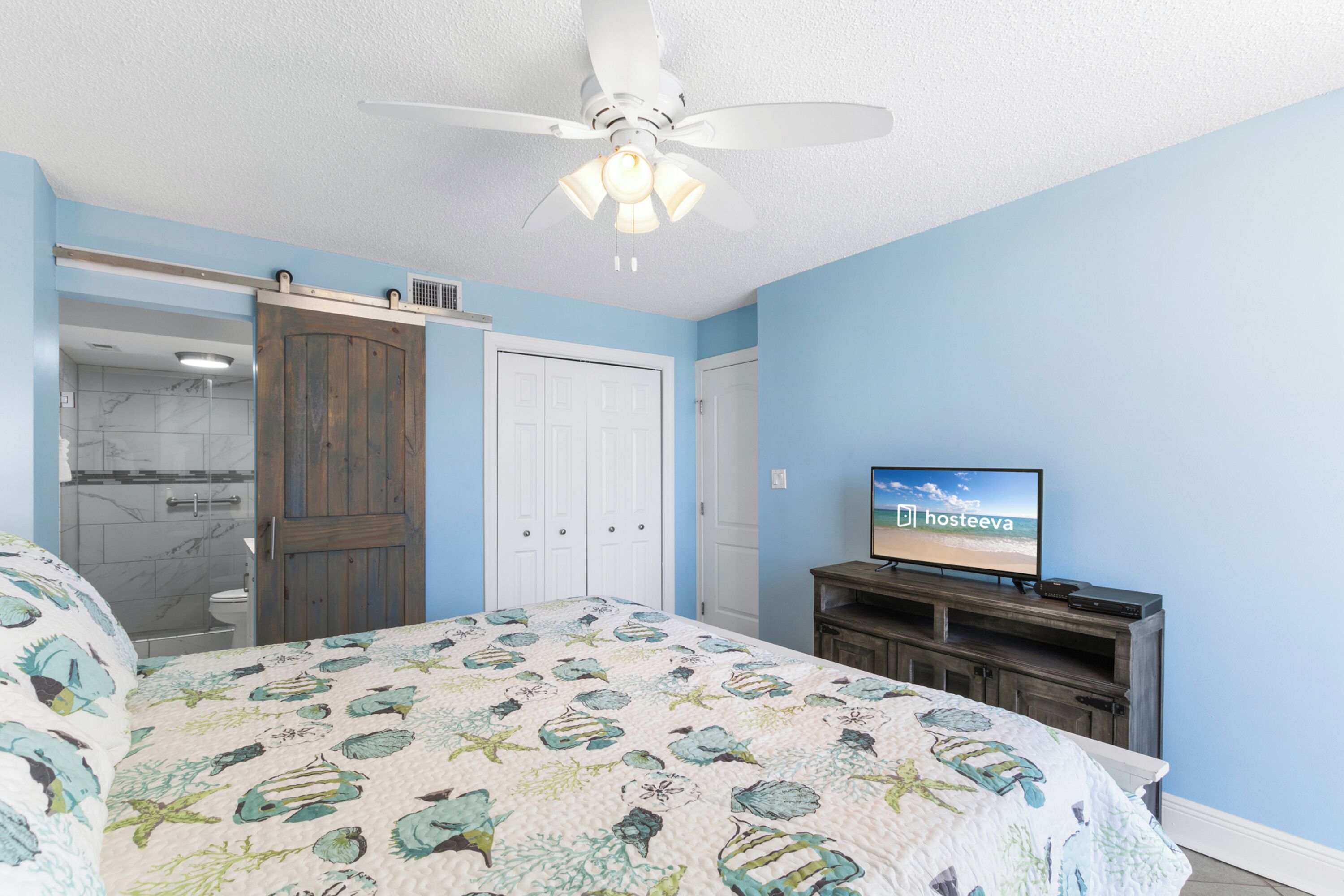 A bedroom with a bed, a ceiling fan, a TV, and a sliding door leading to a bathroom.