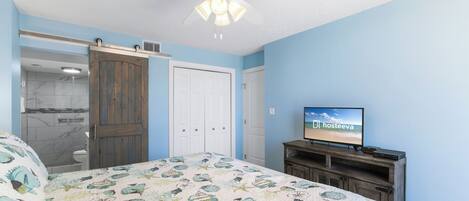 A bedroom with a bed, a ceiling fan, a TV, and a sliding door leading to a bathroom.