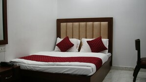 A neatly made bed with a wooden headboard and two red pillows, a bedside table with a phone, and a chair in a simple room with white walls and tiled flooring. - National Lodge (New Delhi)