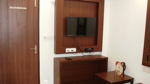 A room with a wooden door, a mounted television, a small wooden table with a remote control, and a small drawer unit. - National Lodge (New Delhi)