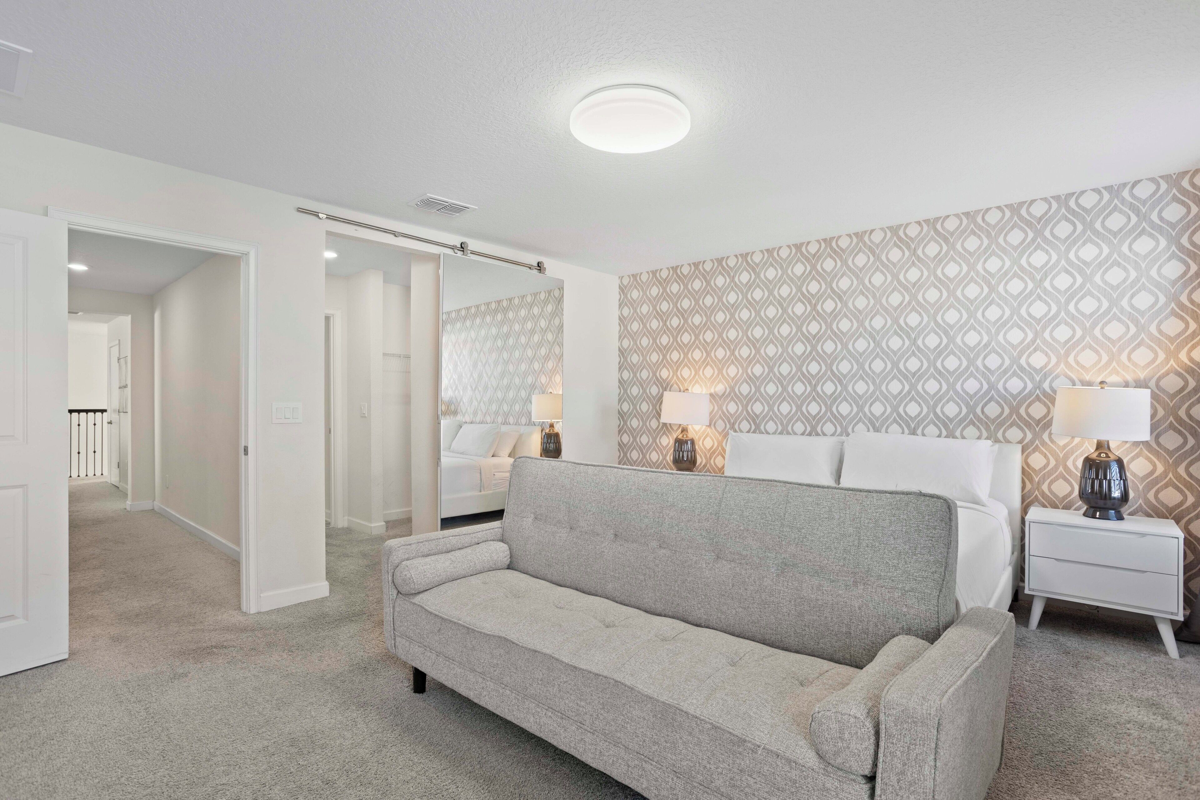 A bedroom with a grey sofa, a white bedside table with a lamp, and a patterned wallpaper on the wall.