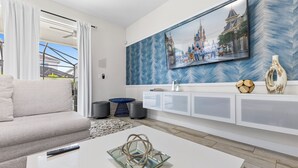 Un soggiorno moderno con un divano, un tavolino da caffè con un oggetto decorativo, una TV montata su una parete azzurra a motivi decorativi e un mobile bianco con un modellino di nave.