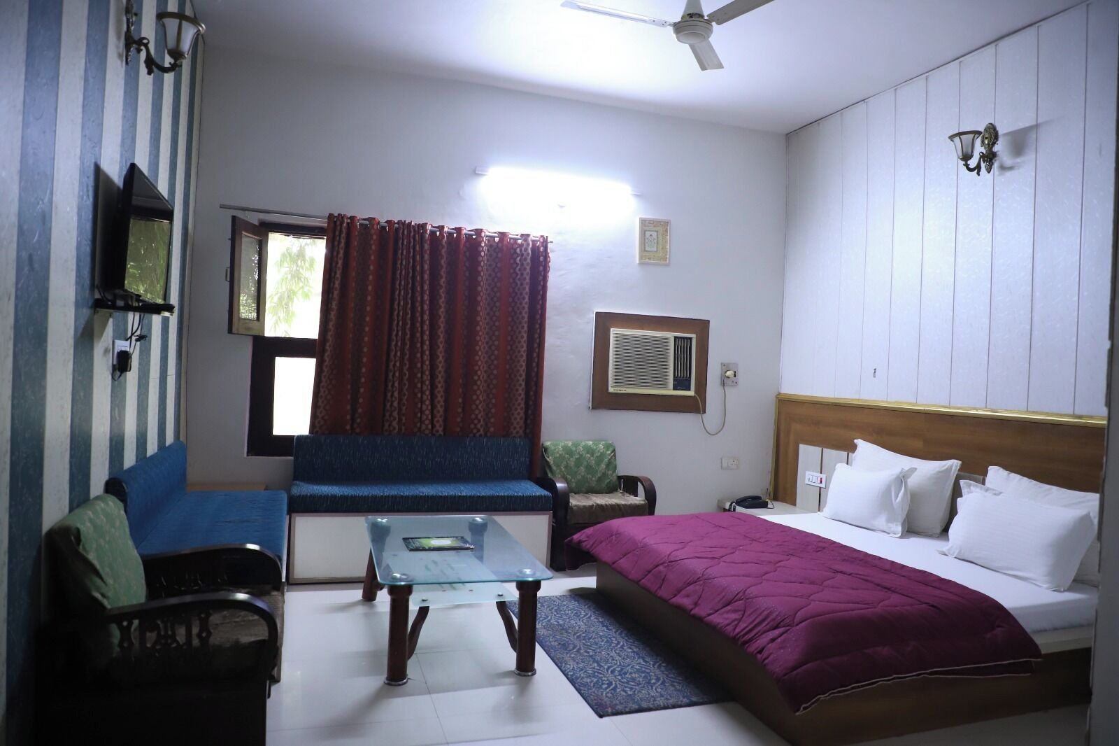 Une chambre d’hôtel comprenant un lit, un canapé, une petite table, une chaise, une fenêtre avec des rideaux, un climatiseur et un ventilateur de plafond.