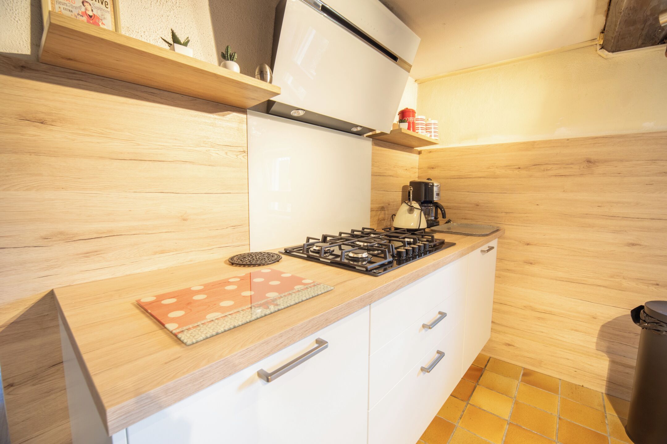 A kitchen with a gas stove, white cabinets, wooden shelves, and a coffee machine.