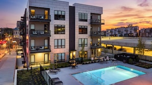 Un hotel moderno de varios pisos con piscina en la azotea, mobiliario exterior y vistas de la ciudad al atardecer.