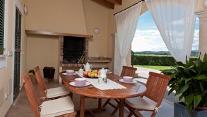 Een terras buiten met een ronde houten tafel, stoelen, een barbecue en uitzicht op het landschap.