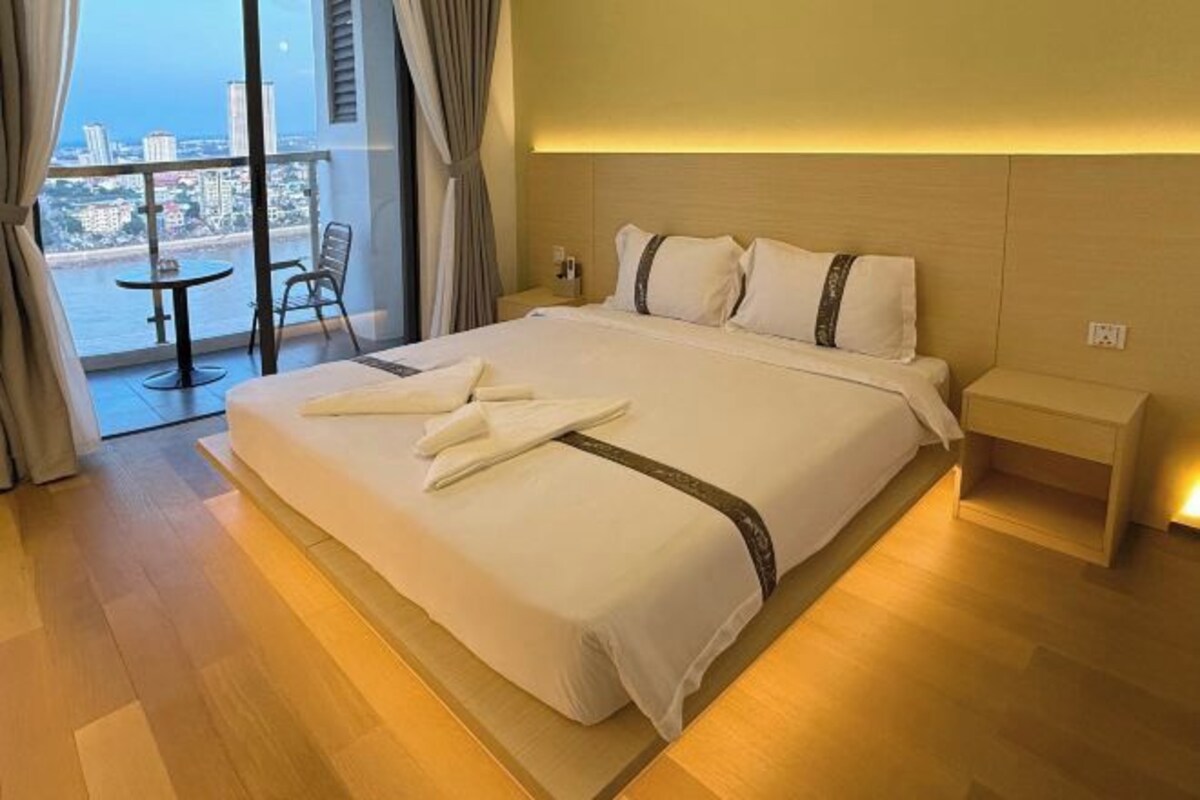 A hotel room with a large bed, a bedside table, a view of the city, and a balcony with a table and chair.