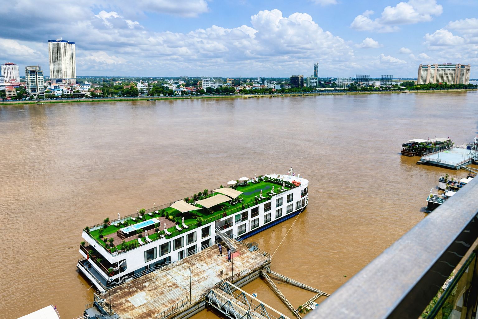 Um barco de passeio com convés verde e assentos externos, atracado em um cais com um horizonte urbano ao fundo.