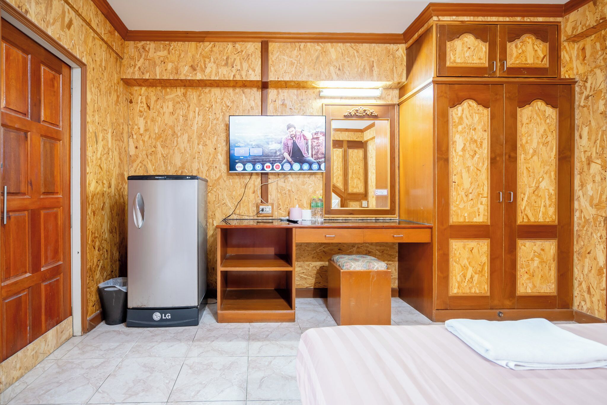 A hotel room with a wooden wardrobe, a desk with a flat-screen TV, a refrigerator, and a bed.