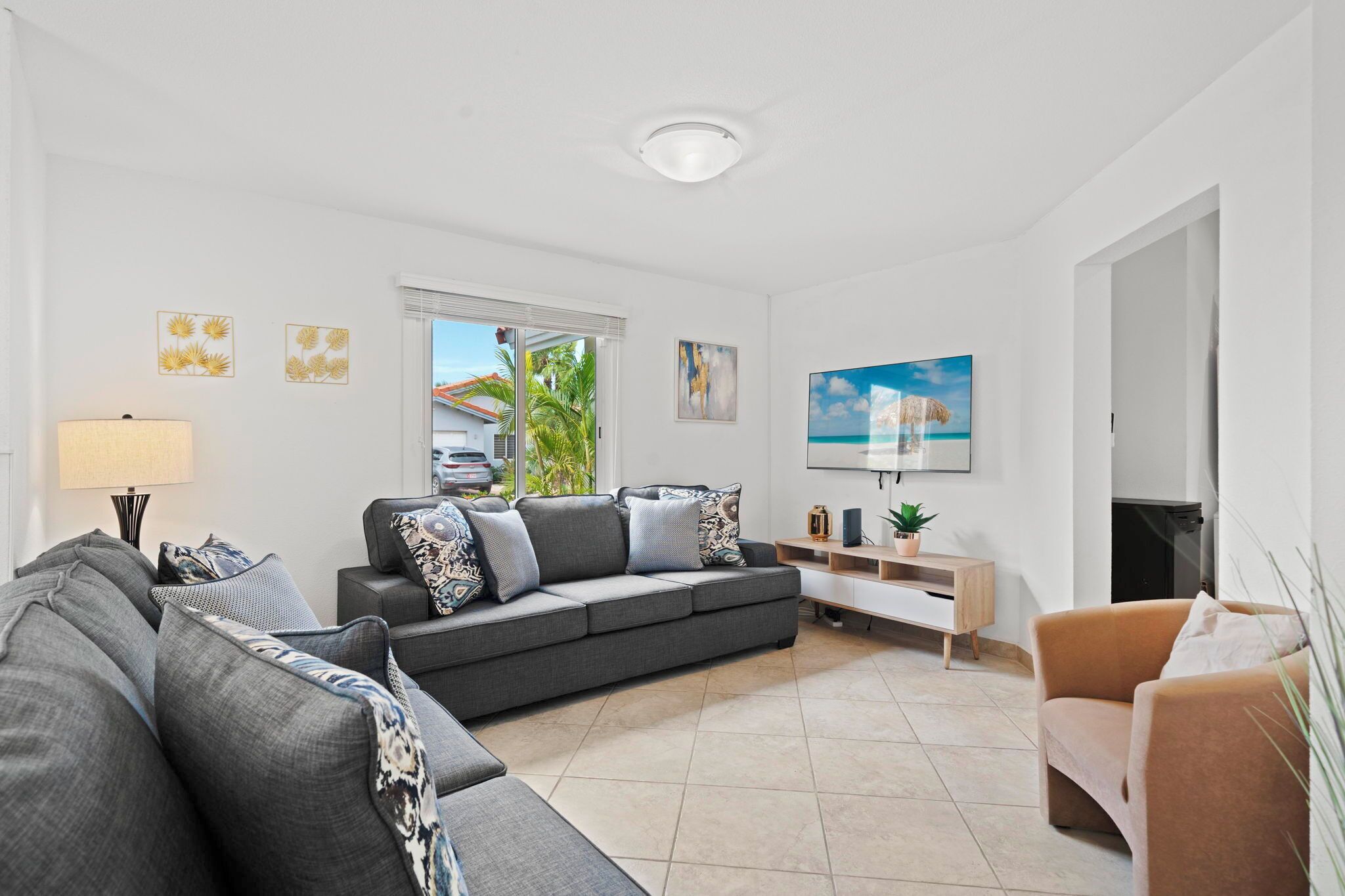 Ein modernes Wohnzimmer mit einem grauen Sofa, einem braunen Sessel und einem Couchtisch. Ein Fenster bietet Blick auf Palmen und ein Haus.