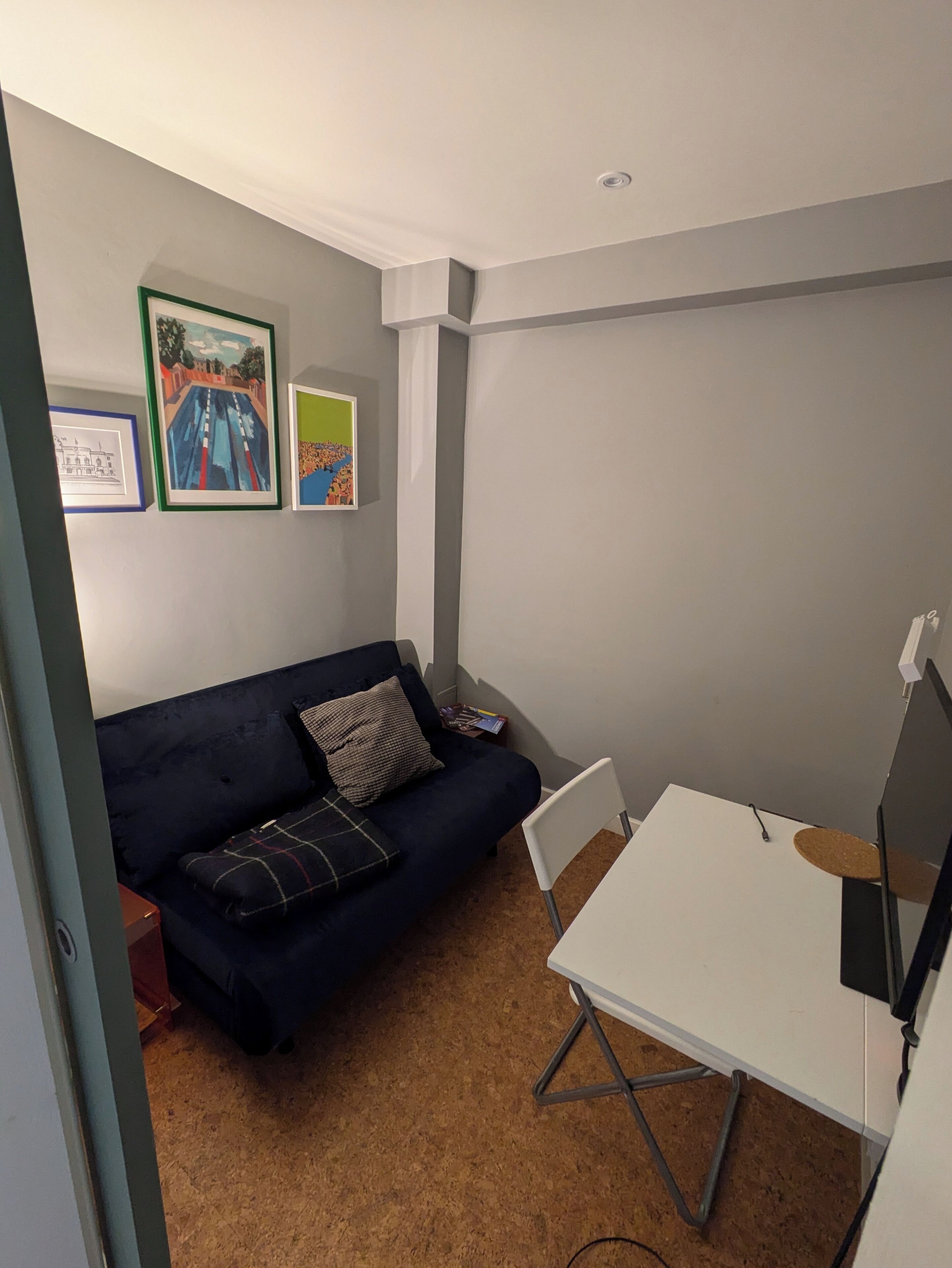 Ein kleiner, schlicht eingerichteter Raum mit einem dunkelblauen Sofa, einem weißen Tisch und drei gerahmten Kunstwerken an der Wand.