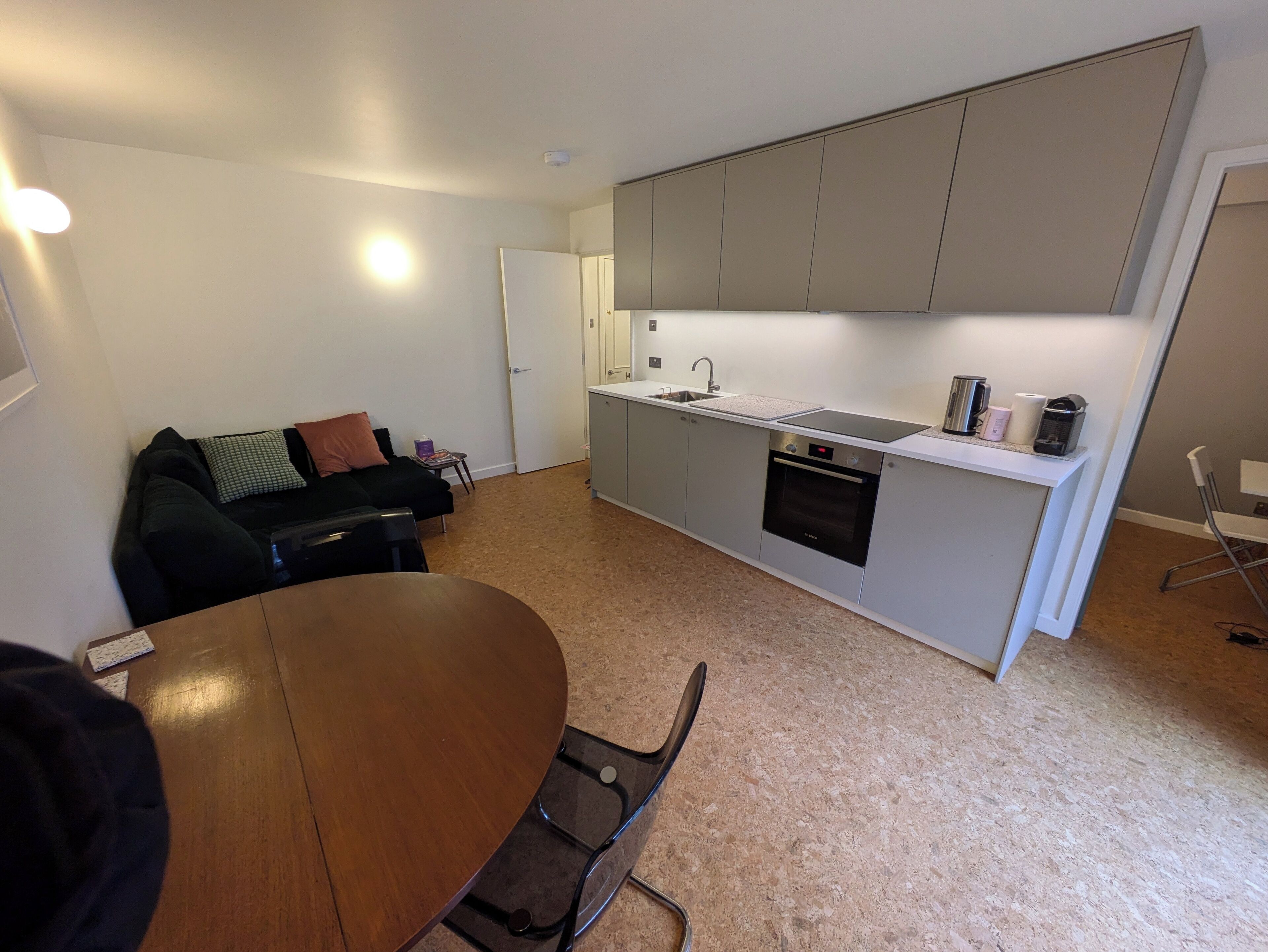 A compact kitchen with grey cabinets, a built-in oven, and a sink. A dining table with chairs is in the foreground.