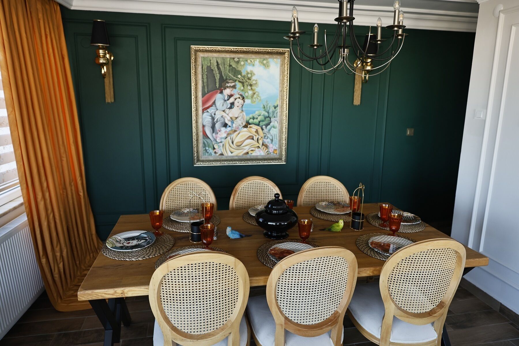 Comedor con mesa de madera dispuesta para seis comensales, rodeada de sillas de mimbre, un candelabro y un cuadro enmarcado en la pared.