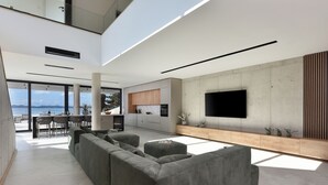 Una sala moderna con un amplio sofá seccional gris, un televisor de pantalla plana montado en una pared de concreto, y un comedor con vista al océano.