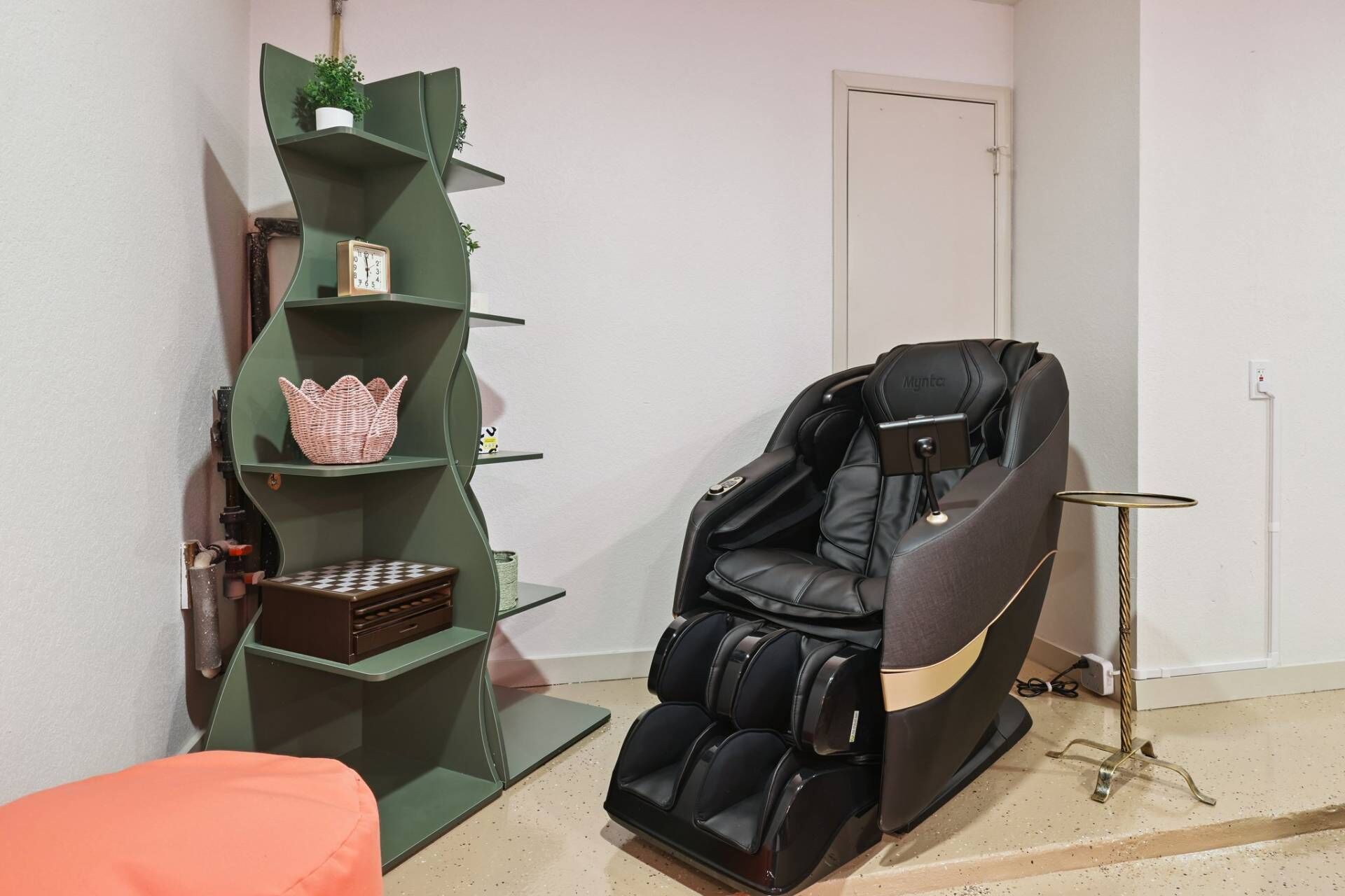A massage chair, a green shelving unit with a clock, and a small table with a plant.
