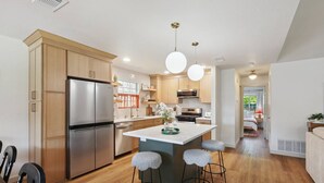 A modern kitchen with wooden cabinets, stainless steel appliances, a central island with stools, pendant lighting, and a view to a bedroom. - Newly Remodeled, Whimsical Retreat -  Viking Hot Tub, Ultimate Game Room, Massage Chair, BBQ (Round Rock)
