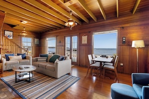 Une pièce avec un plafond en bois, un canapé, une table à manger avec des chaises et une vue sur la mer grâce à de grandes fenêtres.