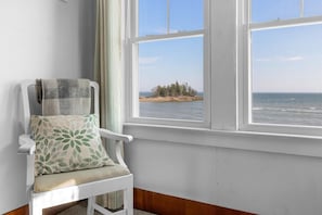 A white chair with a patterned cushion, a window with a view of a beach and trees, and a wooden floor. - The Spruces: Classic 5BR ocean-views in Owls Head with deck, grill, and space for 10 (Owls Head)