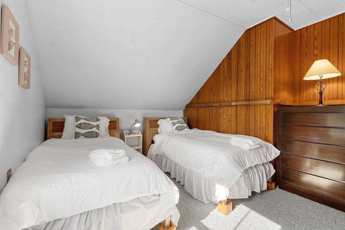 A bedroom with two beds, wooden paneling on the walls, a nightstand with a lamp, and a wooden wardrobe.