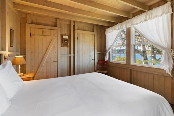 A wooden cabin-style room with a bed, a nightstand, a lamp, a window with curtains, and a door.