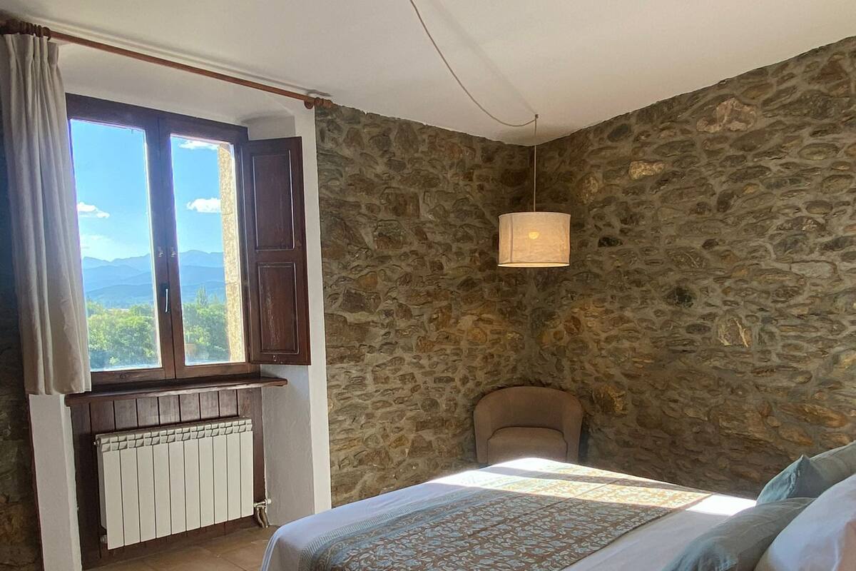 Une chambre avec un grand lit, un mur en pierre, une fenêtre avec des rideaux, un radiateur et une chaise.