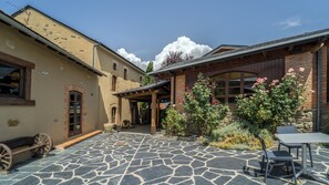 Un patio con suelo de piedra, una mesa de madera y sillas. Se observan edificios con fachadas de ladrillo y enlucido, además de una pérgola de madera.