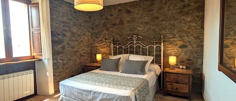 Un dormitorio con pared de piedra, una cama con cabecero metálico, una mesita de noche con una lámpara y una ventana con cortinas.