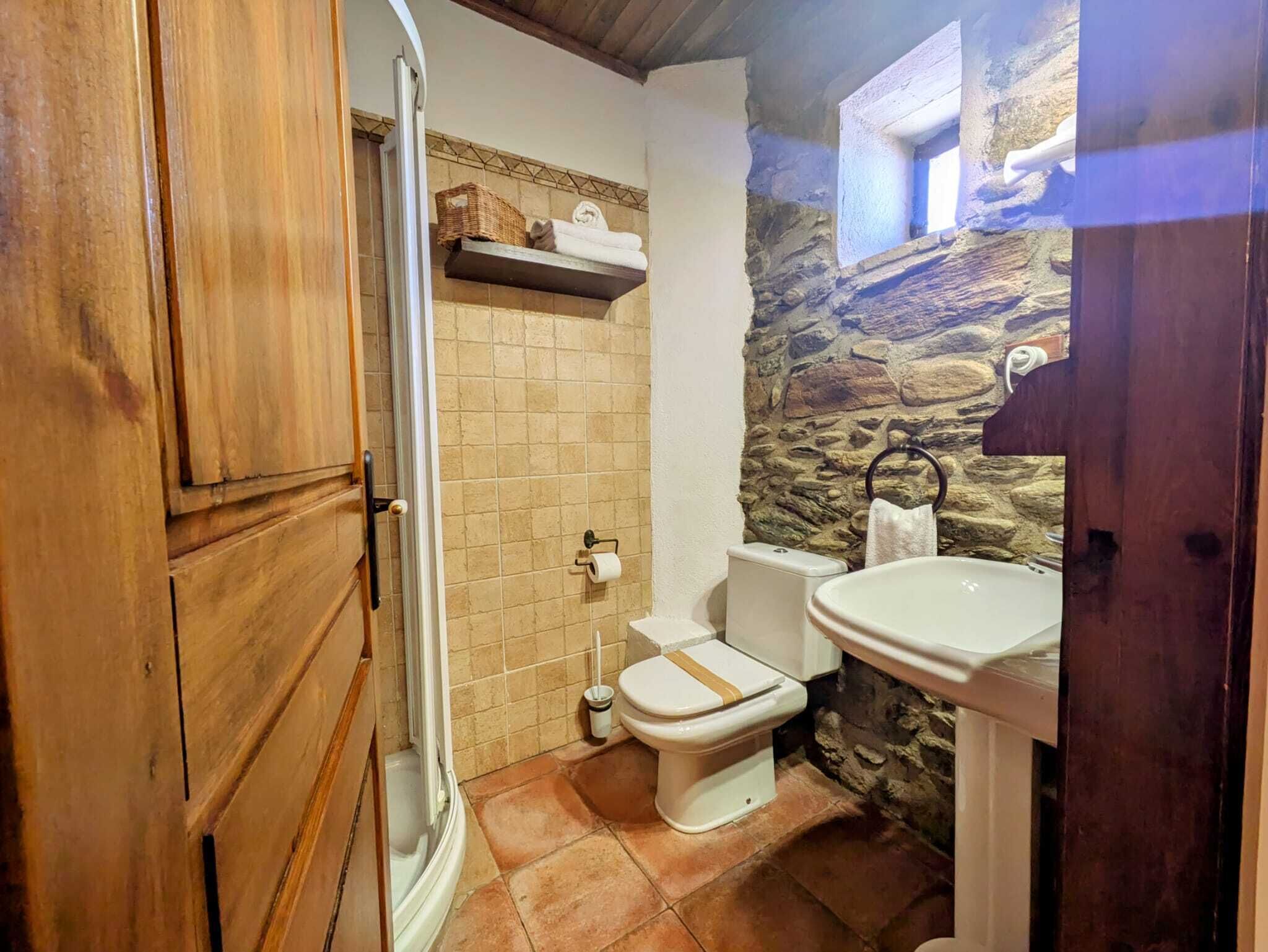 A bathroom with a stone wall, a wooden door, a toilet, a sink, and a shelf with towels.